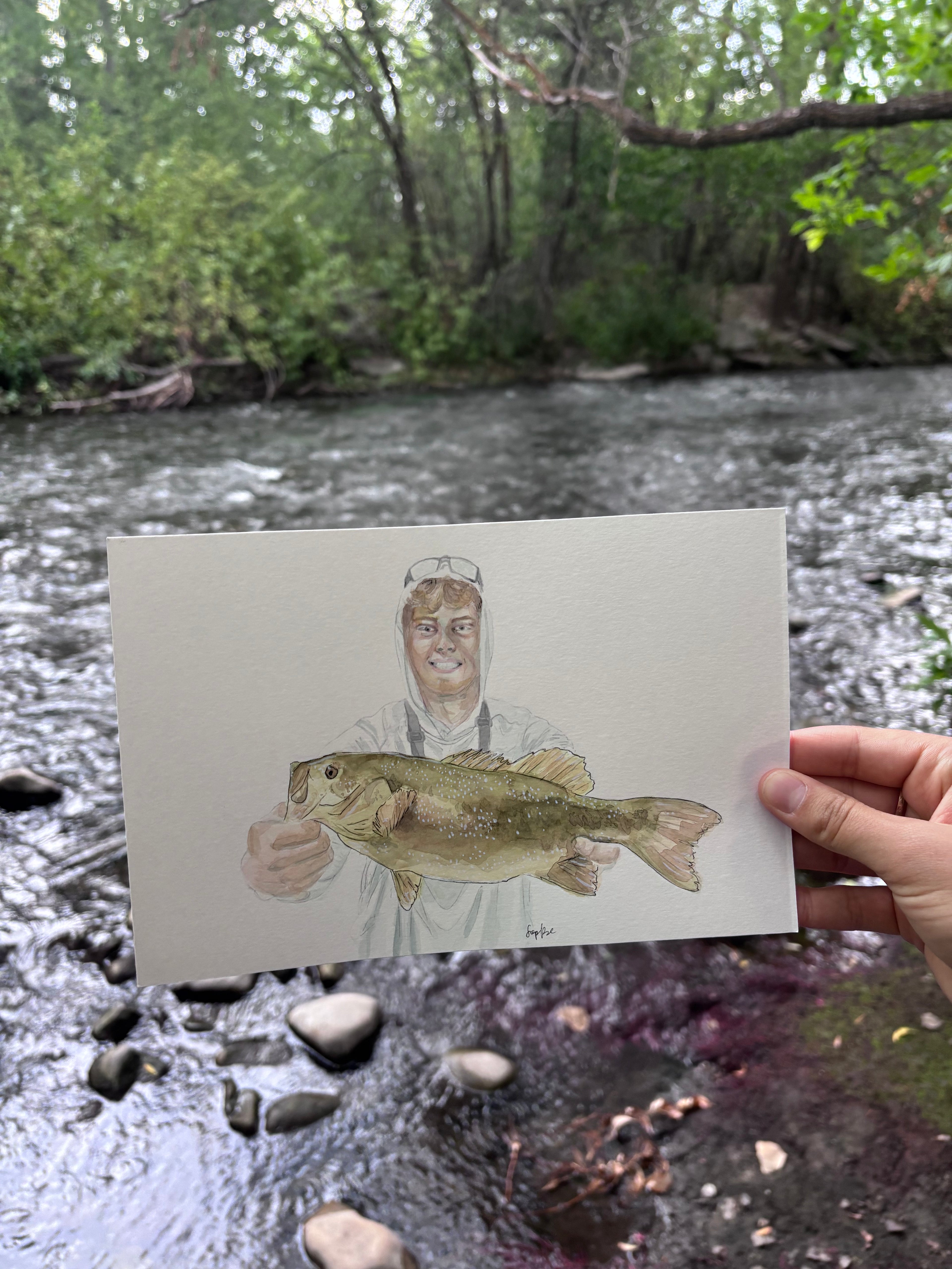 A hand holds a painted art card showing a painted fisherman holding a smallmouth bass, set against a real outdoor river scene with rocks and green trees.