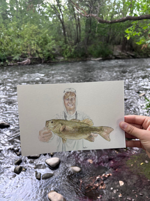 A hand holds a painted art card showing a painted fisherman holding a smallmouth bass, set against a real outdoor river scene with rocks and green trees.