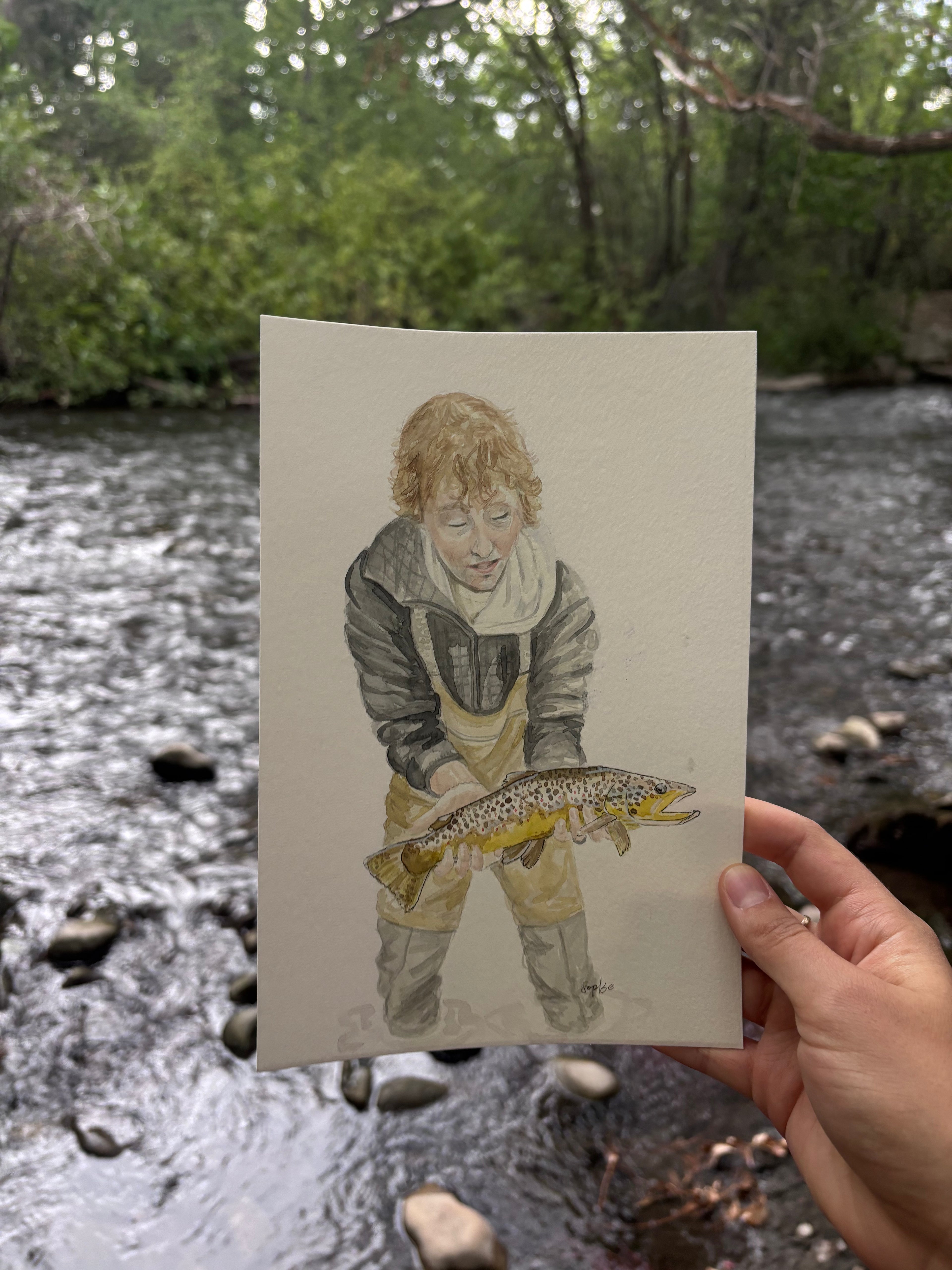 A hand holds a vertical art painting depicting a young boy holding a trout, set against a blurred outdoor background of a flowing river and green trees.